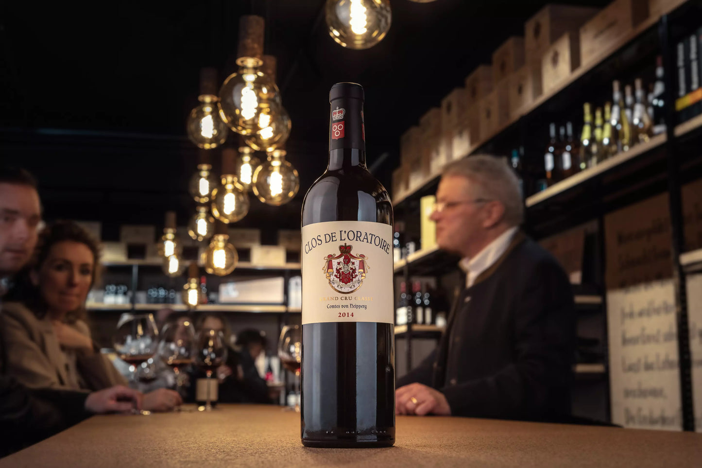 Flasche Clos de l'Oratoire auf dem Tisch in der Vinothek Hauser mit Gästen im Hintergrund am Tisch.