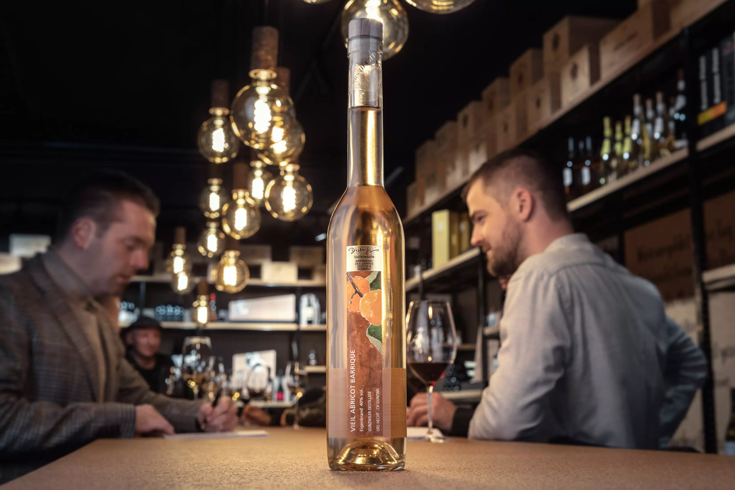 Vieil Abricot von Gunzwiler Destillate Urs Hecht auf dem Tisch in der Vinothek Hauser mit Gästen im Hintergrund.