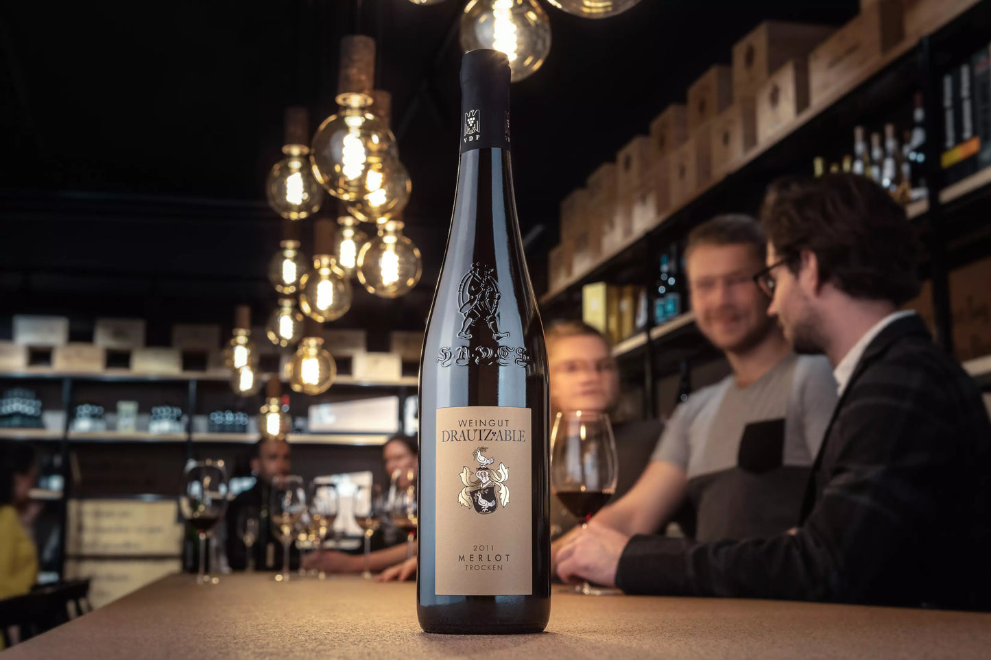 Flasche Merlot HADES vom Weingut Drautz-Able auf dem Tisch in der Vinothek Hauser mit Gästen im Hintergrund.