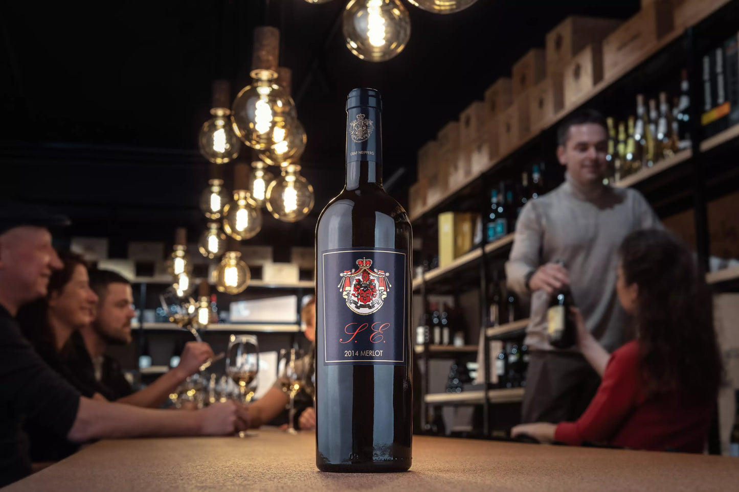Vinothek Hauser mit vielen Gästen beim Apéro, im Vordergrund eine Flasche Merlot S.E. vom Weingut Graf Neipperg präsentiert im Fokus auf dem Tisch.