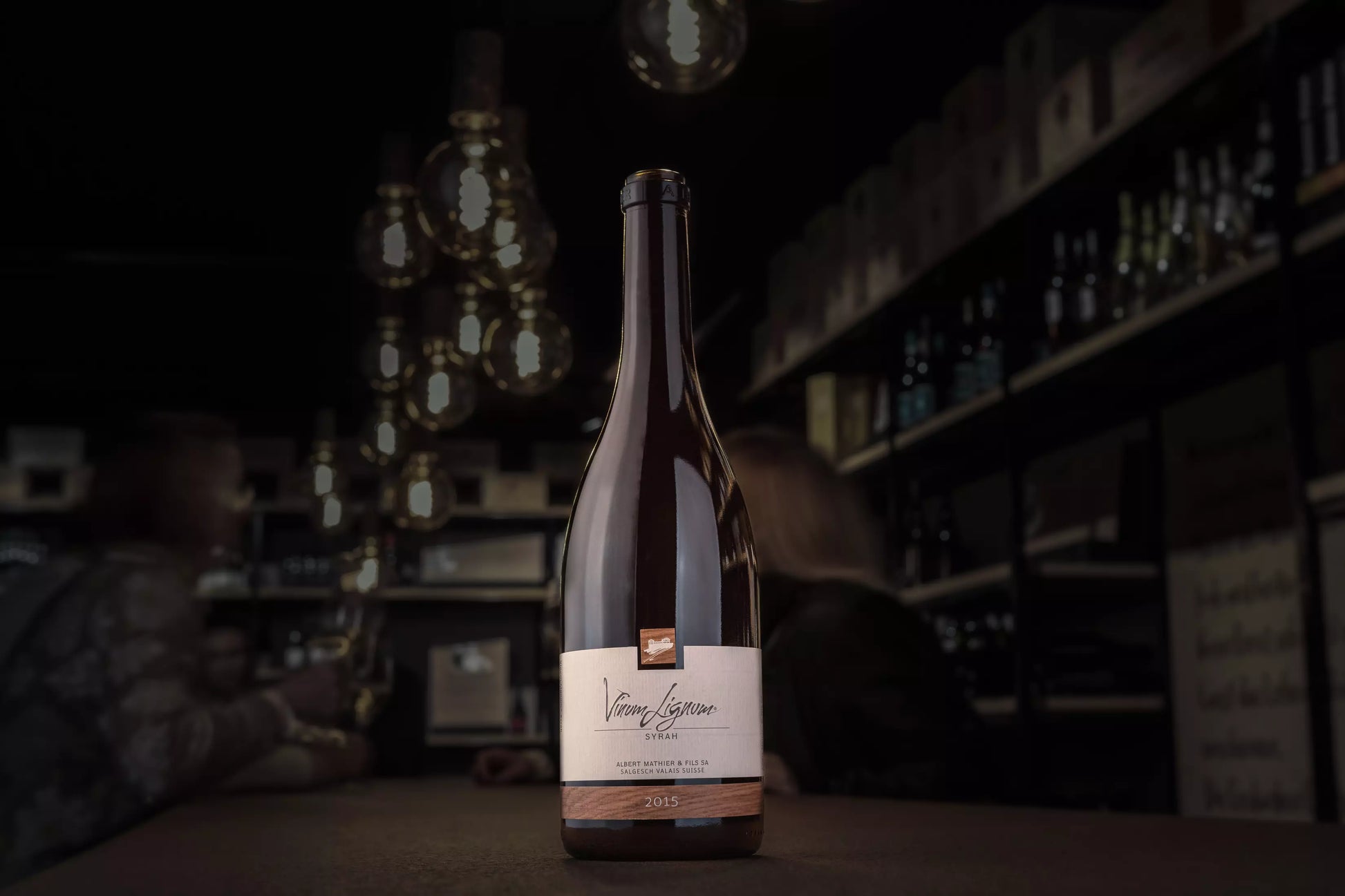 Dunkle Vinothek Hauser mit einer Flasche Vinum Lignum Syrah auf dem Tisch im Vordergrund.