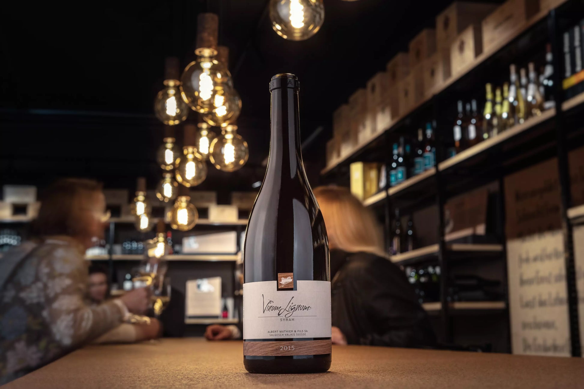 Flasche Vinum Lignum Syrah auf dem Tisch in der Vinothek Hauser mit Personen im Hintergrund.
