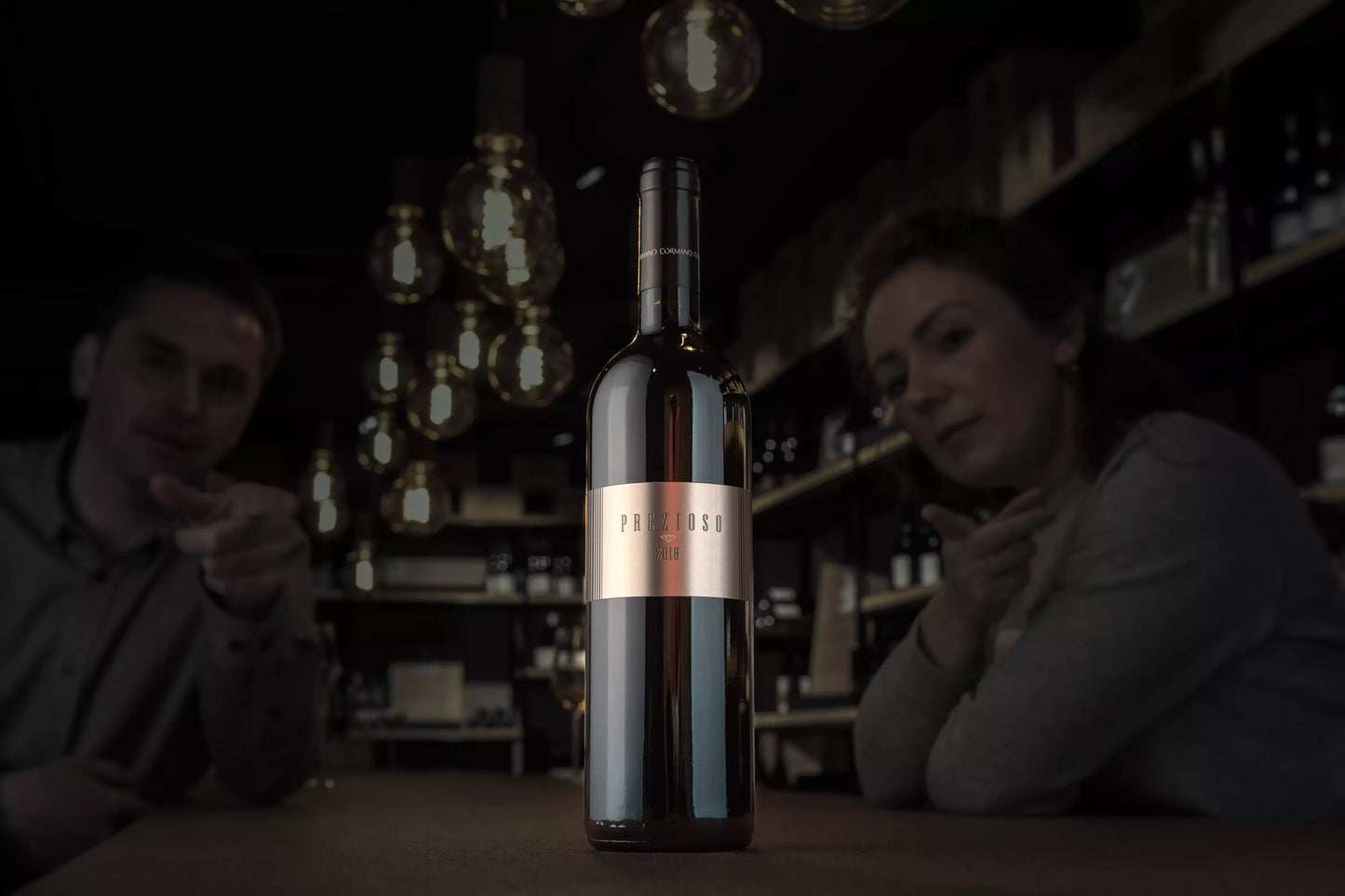 Dunkle Vinothek, im Mittelpunkt eine beleuchtete Flasche Prezioso Rosso del Ticino von Cormano Vini auf dem Tisch.