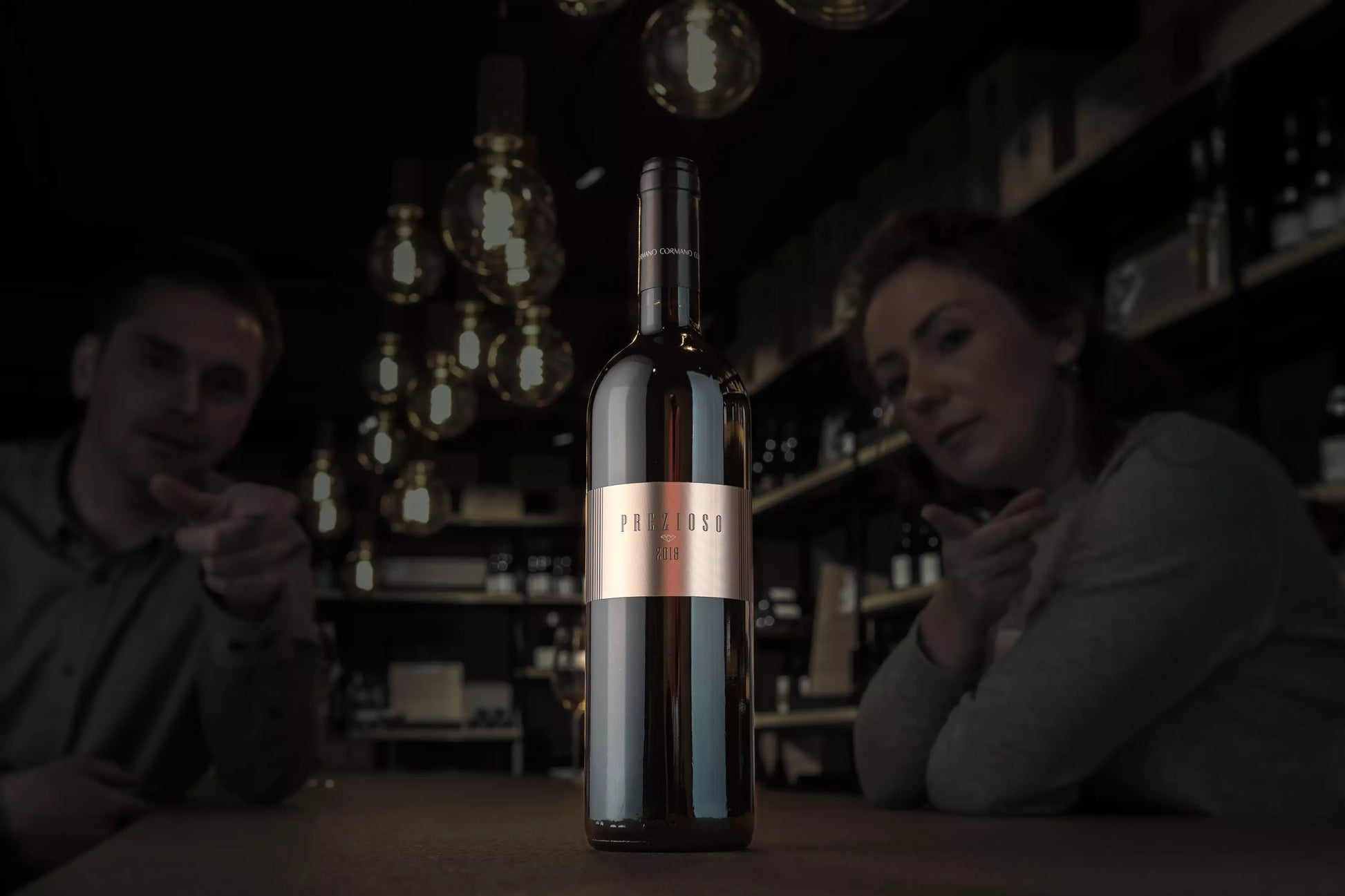 Dunkle Vinothek, im Mittelpunkt eine beleuchtete Flasche Prezioso Rosso del Ticino von Cormano Vini auf dem Tisch.