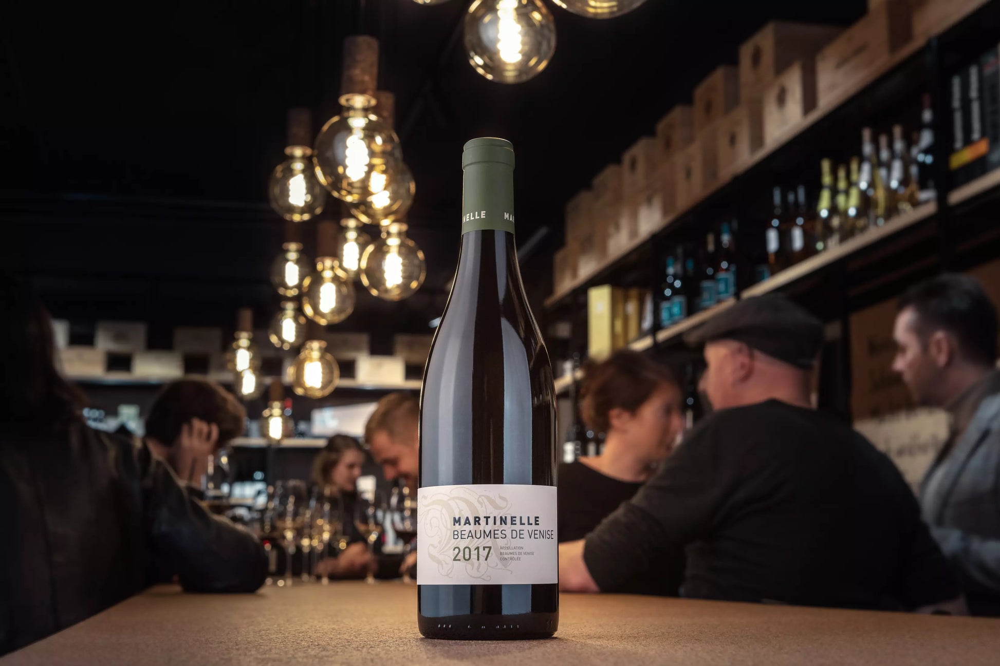 Beaumes de Venise von der Domaine Martinelle auf dem Tisch in der Vinothek Hauser mit Gästen beim Apéro im Hintergrund.