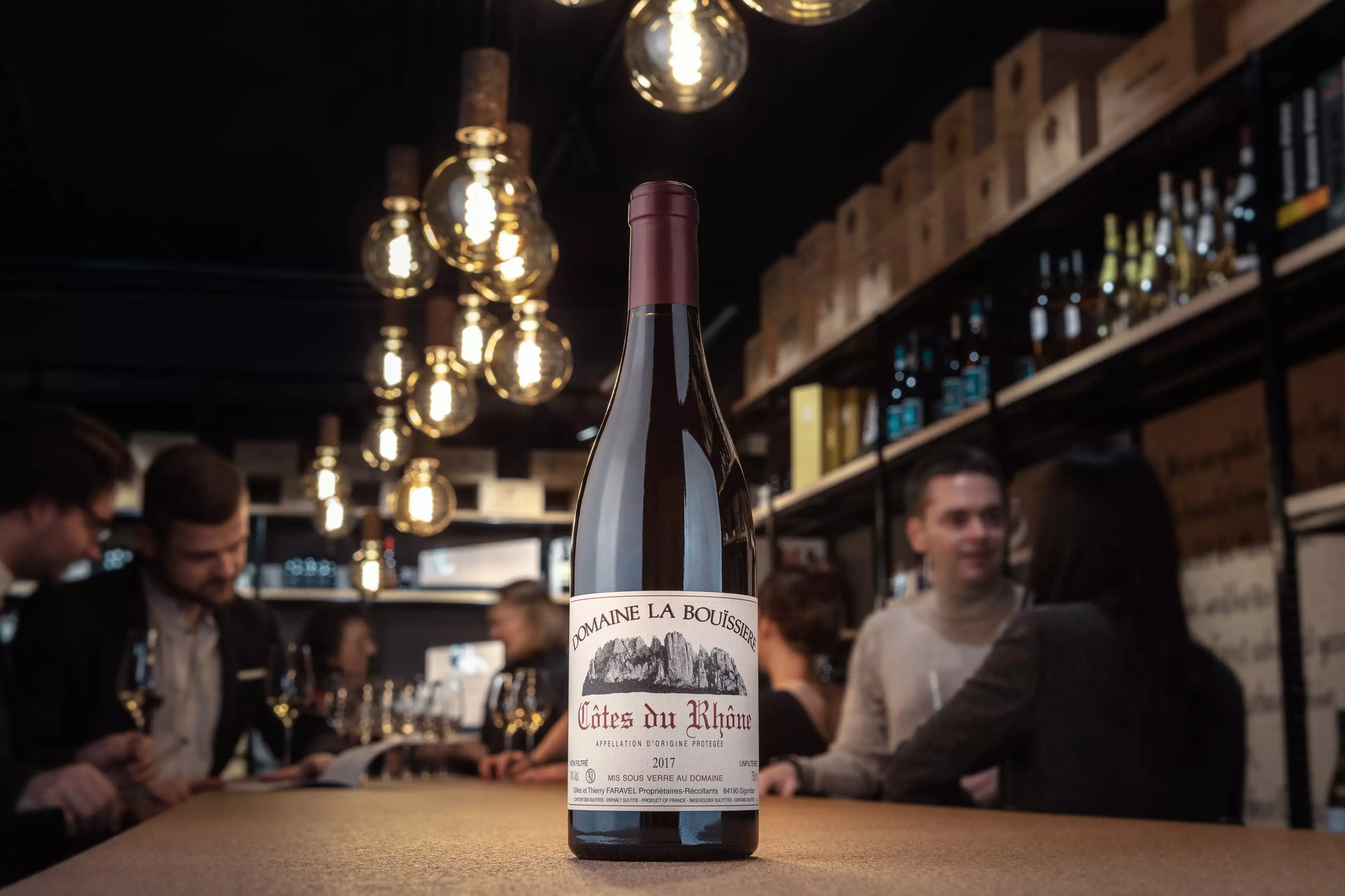 Côtes du Rhône von der Domaine La Bouissière auf dem Tisch in der Vinothek Hauser mit Gästen beim Apéro im Hintergrund.