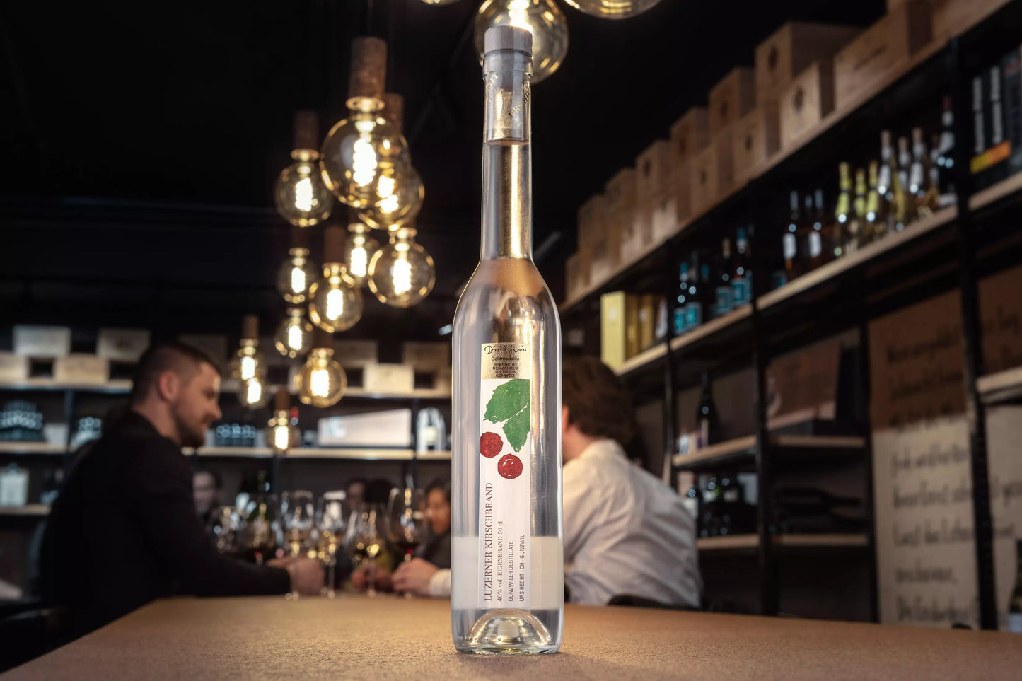 Luzerner Kirsch von Gunzwiler Destillate Urs Hecht auf dem Tisch in der Vinothek Hauser mit Gästen im Hintergrund.