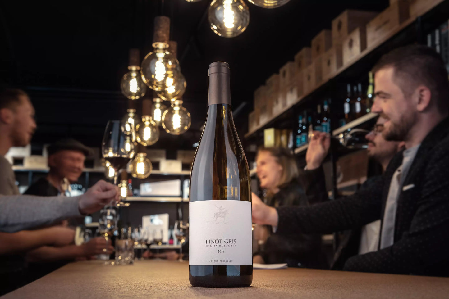 Pinot Gris vom Johanniterkeller Martin Hubacher auf dem Tisch in der Vinothe kHauser mit Gästen im Hintergrund.