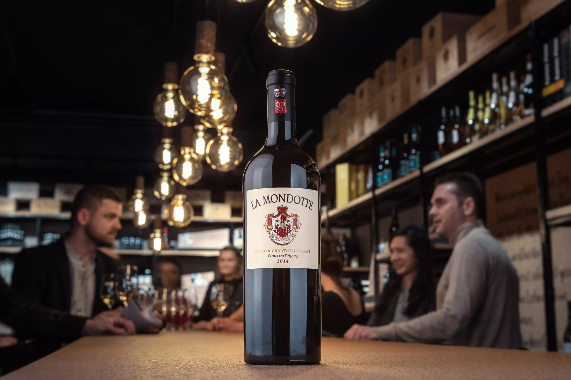 La Mondotte Premier Grand Cru Classé auf dem Tisch in der Vinothek Hauser mit Gästen im Hintergrund.
