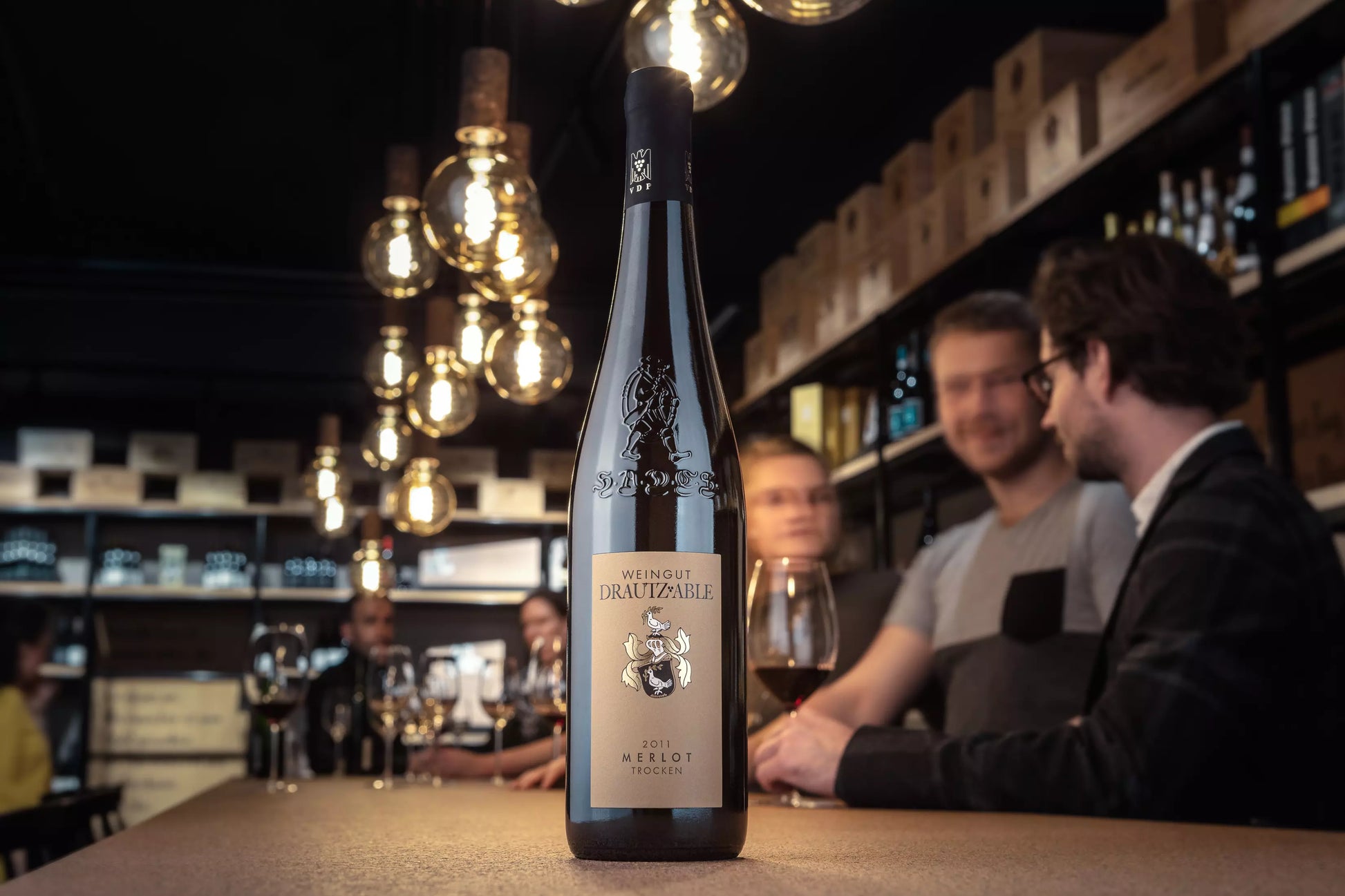 Flasche Merlot HADES vom Weingut Drautz-Able auf dem Tisch in der Vinothek Hauser mit Gästen im Hintergrund.