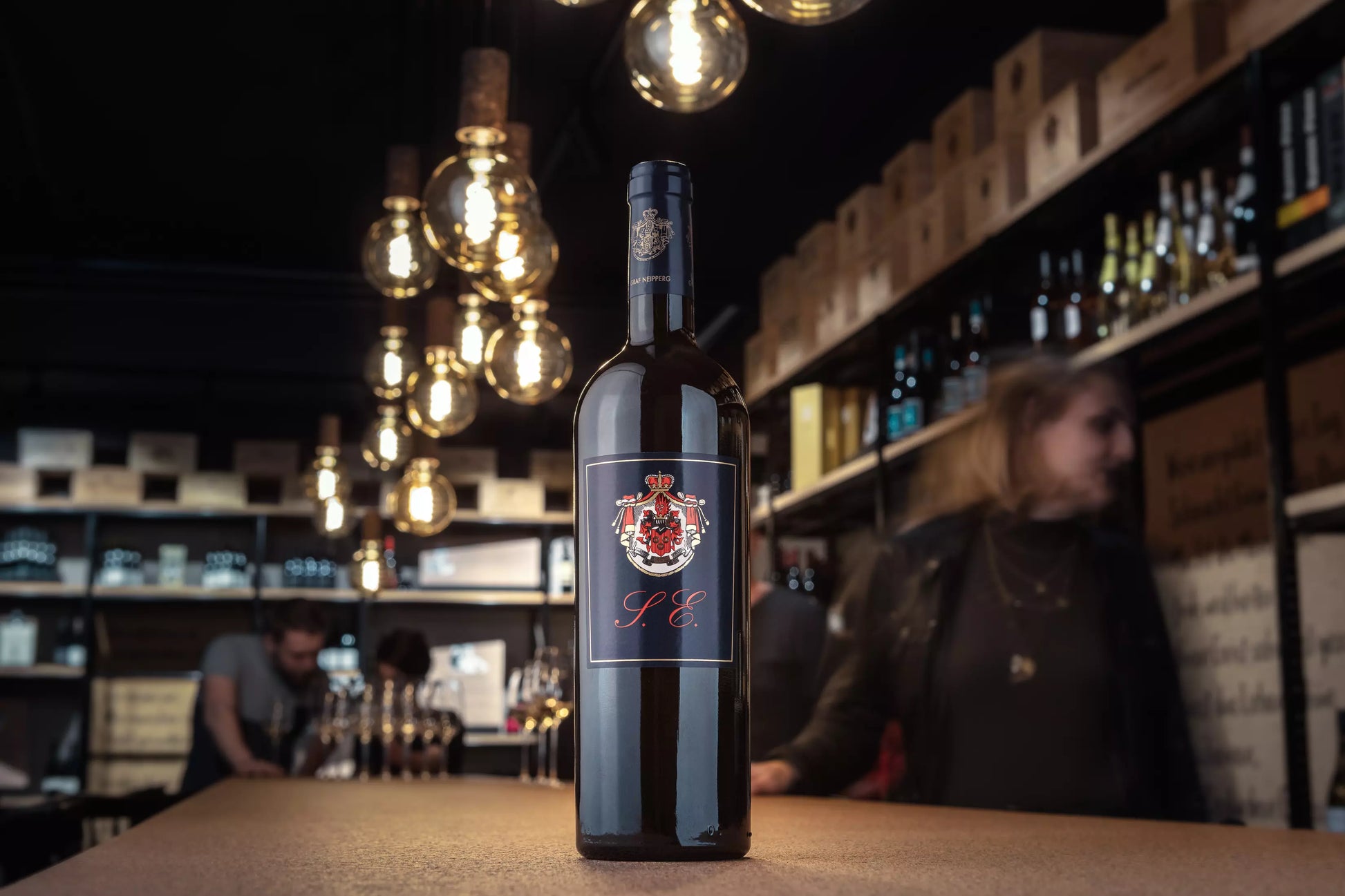 Flasche Cuvéee S.E vom Weingut Graf Neipperg in der Vinothek Hauser auf dem Tisch mit Gästen im Hintergrund.
