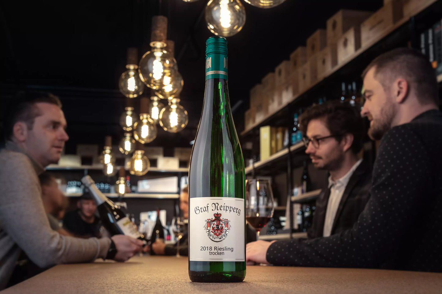Flasche Riesling VDP.Gutswein vom Weingut Graf Neipperg auf dem Tisch in der Vinothek Hauser mit Gästen im Hintergrund.