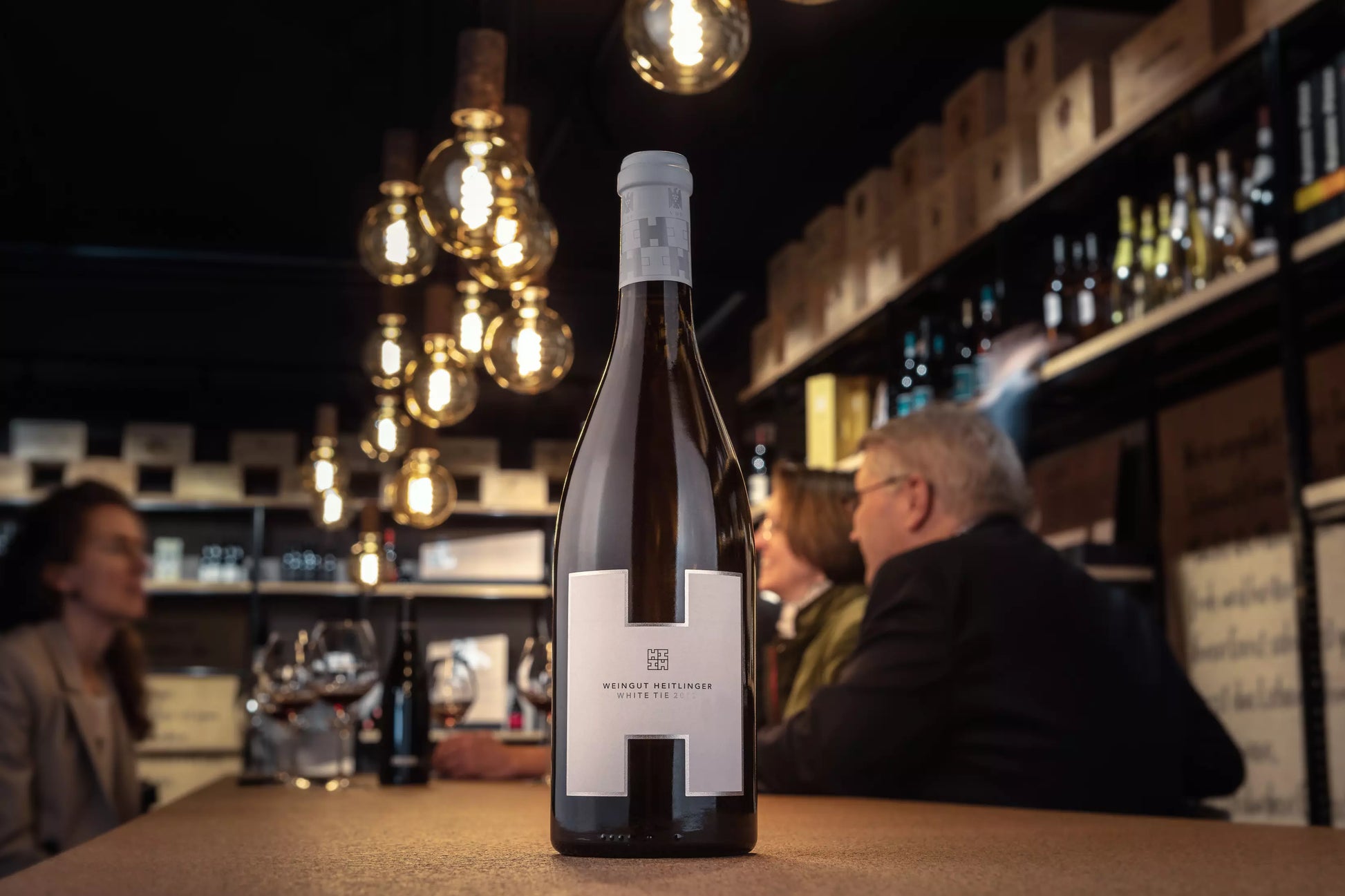 Flasche White Tie vom Weingut Heitlinger auf dem Tisch in der Vinothek Hauser mit Gästen im Hintergrund.