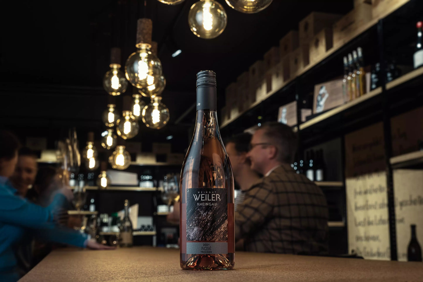 Flasche Rosé trocken vom Weingut Weiler auf dem Tisch in der Vinothek Hauser mit Gästen im Hintergrund.