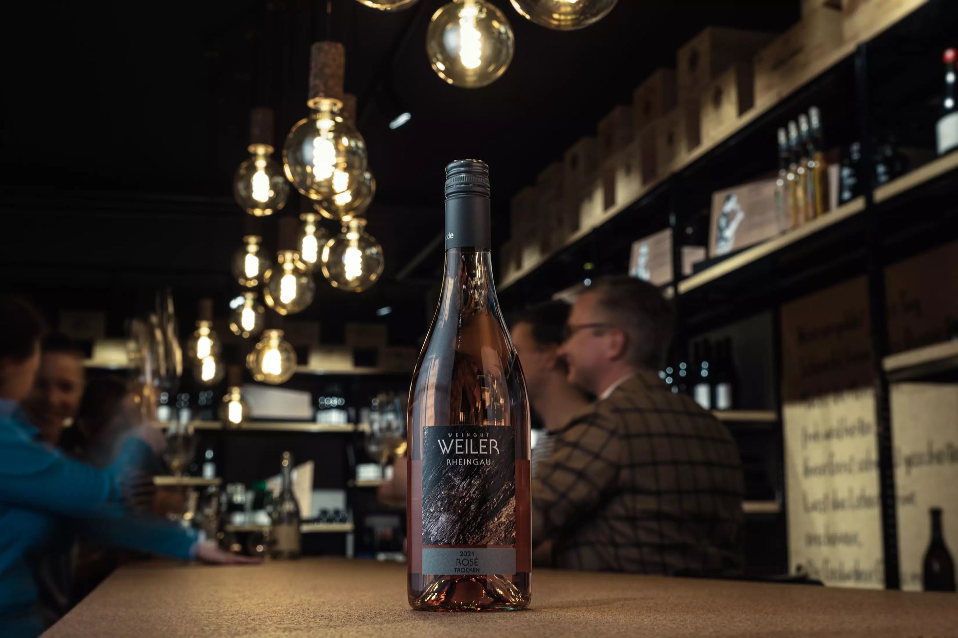 Flasche Rosé trocken vom Weingut Weiler auf dem Tisch in der Vinothek Hauser mit Gästen im Hintergrund.