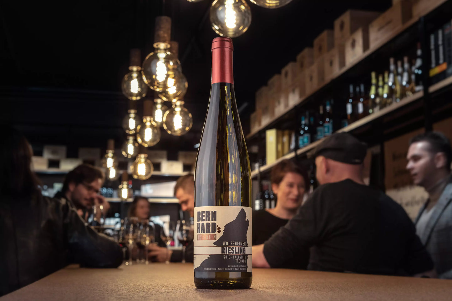 Flasche Wolfsheimer Riesling vom Weingut Bernhard in der Vinothek Hauser auf dem Tisch mit Gästen im Hintergrund.